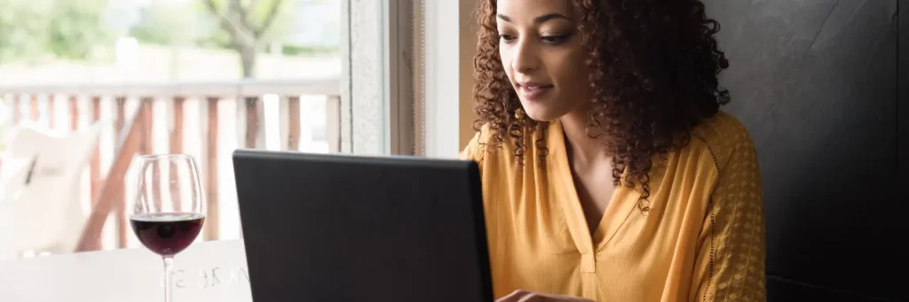 Woman in a yellow blouse working on a laptop indoors next to a window with a glass of red wine on the table.